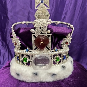 May include: A bejeweled crown with a large red gemstone at its center. The crown features a silver frame adorned with pearls, clear and colored gemstones, and a white fur trim. It sits atop a purple velvet cushion.