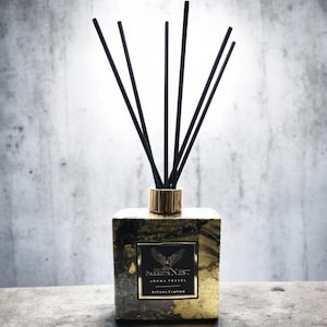 Puede incluir: Botella difusora negra y dorada con cañas negras y una tapa dorada. La botella tiene un diseño marmoleado negro y dorado y el texto "The Parrot's Nest Aroma Vessel Artisan Crafted".