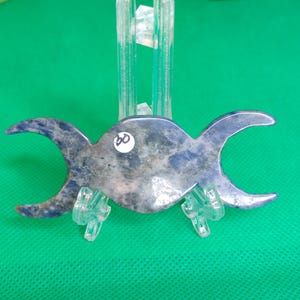 Puede incluir: Un colgante de triple luna azul y blanco hecho de lapisl&aacute;zuli, exhibido en un soporte de pl&aacute;stico transparente. El colgante tiene un di&aacute;metro de aproximadamente 7,5 cm.
