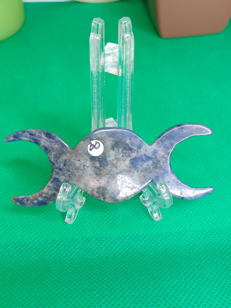 Puede incluir: Un colgante de triple luna azul y blanco hecho de lapisl&aacute;zuli, exhibido en un soporte de pl&aacute;stico transparente. El colgante tiene un di&aacute;metro de aproximadamente 7,5 cm.