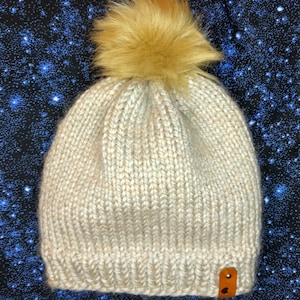 May include: A knitted beanie in a cream colour, topped with a fluffy, light brown pom-pom. The hat features a ribbed knit pattern and a small, brown leather tag. The background is a dark blue fabric with white star-like patterns.