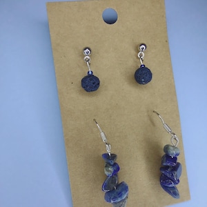 May include: Two pairs of earrings displayed on a brown card. One pair features dark blue, textured spheres suspended from silver-tone hooks. The other pair showcases a cascade of blue and gray gemstone chips on silver-tone hooks. Jewelry.