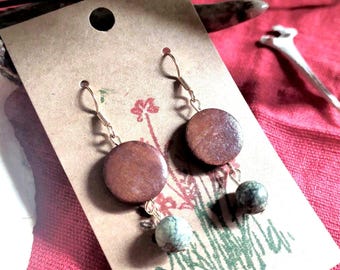Peut inclure: Une paire de boucles d'oreilles pendantes composée d'un disque en bois brun circulaire suspendu à un crochet en métal doré, avec une petite perle en pierre sphérique marbrée vert et gris accrochée au bas de chaque disque par un fil métallique.