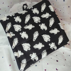 May include: Black and white quilted pot holder with a loop for hanging. The pot holder features a repeating pattern of white ghost illustrations on a black background. The edges are neatly sewn, and the overall design is square.