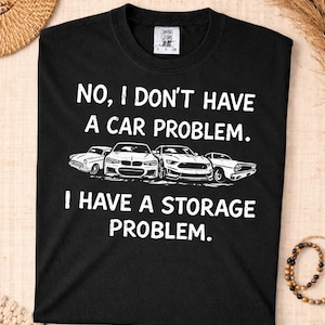 Può includere: T-shirt nero con testo bianco: "NO, I DON'T HAVE A CAR PROBLEM. I HAVE A STORAGE PROBLEM." Il design include disegni a tratto di quattro auto.