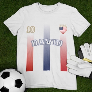 May include: White football shirt with the name "DAVID" in blue, the number "10" in gold, and a USA flag emblem. Red and blue stripes run vertically down the front. A football and white gloves are in the image.