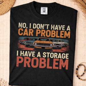 Puede incluir: Camiseta negra con el texto "NO, I DON'T HAVE A CAR PROBLEM I HAVE A STORAGE PROBLEM" en blanco y naranja. El gráfico presenta coches clásicos de varios colores. La camiseta es de material suave.