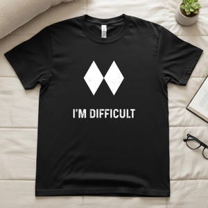 May include: Black t-shirt featuring a white diamond design and the words "I'M DIFFICULT". This casual tee is made from a soft fabric. The graphic is a simple, bold statement. Perfect for everyday wear.