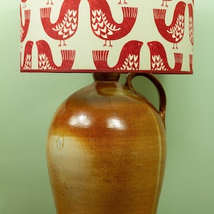 May include: A table lamp with a ceramic jug base and a patterned lampshade. The lampshade has a cream background with a repeating pattern of red birds. The jug is brown with a handle and a glossy finish.