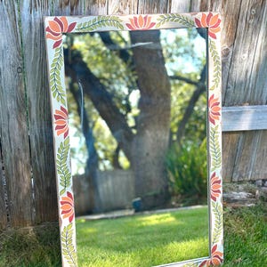 May include: Rectangular mirror with a cream-colored frame adorned with orange and red floral designs and green leaf accents. The mirror is leaning against a wooden fence on a grassy lawn, reflecting the surrounding trees and sky.