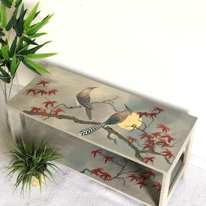 May include: A two-tiered, rectangular wooden table with a hand-painted design. The artwork features two birds perched on a branch with red leaves and a yellow moon against a gray and blue background. The table has a light gray frame.