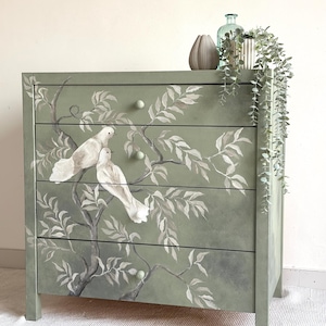 Op de afbeelding: Een saliegroene dressoir met vier lades, versierd met een handgeschilderd ontwerp van witte duiven op takken met grijze en witte bladeren. De dressoir heeft lichtgroene knoppen en is versierd met een vaas, een fles en een plant.