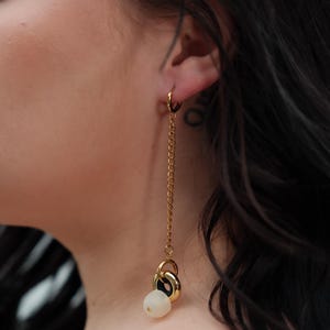 May include: Gold chain earrings with a white bead and gold hoops. The earrings are dangling from the earlobe.