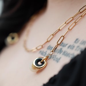 May include: Gold necklace with a pendant comprising a black ring and a white, rounded centre. The chain has elongated links. The necklace is set against a dark background.