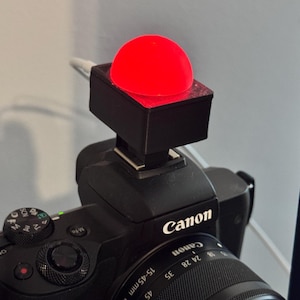 Peut inclure: Un appareil photo noir avec une lumière rouge fixée sur le dessus. L'appareil photo porte le logo Canon et un objectif avec un zoom 15-45 mm.