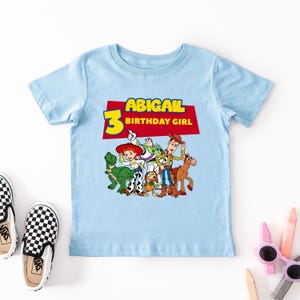 May include: Light blue t-shirt with a red, yellow, and blue graphic of characters from the Toy Story movie franchise. The text "ABIGAIL 3 BIRTHDAY GIRL" is printed on the shirt.