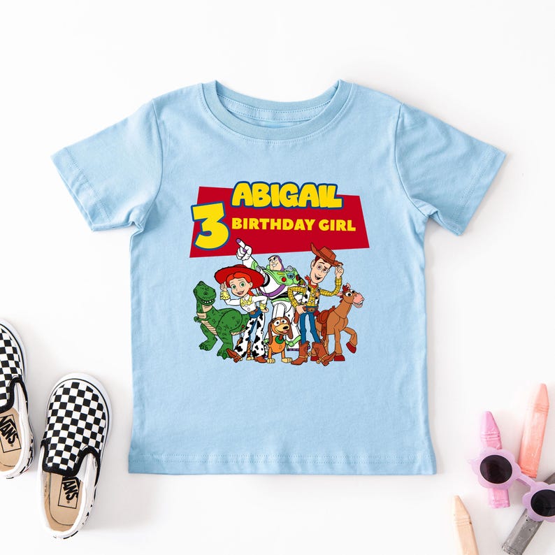 May include: Light blue t-shirt with a red, yellow, and blue graphic of characters from the Toy Story movie franchise. The text "ABIGAIL 3 BIRTHDAY GIRL" is printed on the shirt.