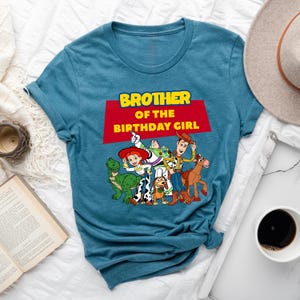 May include: A teal blue t-shirt with a red and yellow graphic of the Toy Story characters. The text "Brother of the Birthday Girl" is printed on the shirt.
