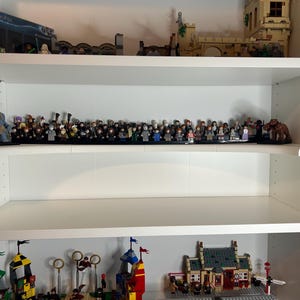 May include: A white shelving unit displays various Lego sets. The top shelf holds a Lego set box and other structures. The middle shelf features a collection of Lego mini-figures. The bottom shelf showcases more Lego structures.