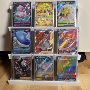 Puede incluir: Un expositor blanco de tres niveles que contiene nueve cartas coleccionables en fundas protectoras transparentes. Las cartas presentan varias ilustraciones coloridas de personajes y criaturas, incluyendo Mew V, Rillaboom, Eldegoss V, Wailord V, Rose, Centiskorch VMAX, Piers, Cursola y Scizor VMAX.