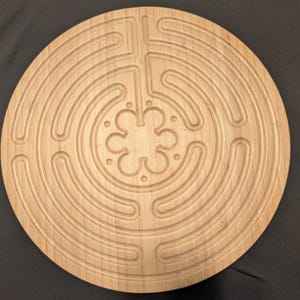 May include: A circular, light brown wooden labyrinth with a carved path design. The labyrinth features a central floral pattern and concentric circular pathways. The wood grain is visible, and the design is centered on the wooden surface.
