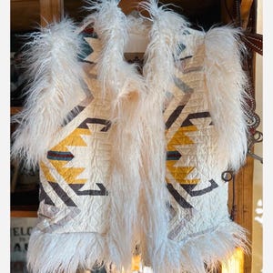 May include: Cream-colored vest with a geometric pattern in yellow, brown, and blue. The vest is trimmed with long, fluffy white faux fur. The design features a quilted texture and a unique, eye-catching style.