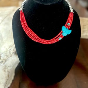 May include: A red beaded necklace with a turquoise butterfly pendant. The necklace features multiple strands of small red beads and silver-coloured end caps. The butterfly pendant is a bright turquoise colour.