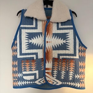 May include: A quilted vest with a geometric pattern in shades of blue, white, and orange. The vest has a cream-colored, fluffy collar and two front pockets. It is hanging on a black hanger.