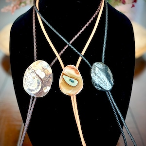 May include: Three bolo ties featuring unique stone pendants. The left pendant is a round, mottled brown and cream stone. The center pendant is a teardrop-shaped tan stone with a green center. The right pendant is a dark gray oval stone with a white leaf pattern.