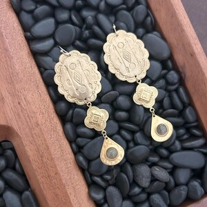 May include: A pair of brass-colored earrings with an oval top, a clover-shaped middle, and a teardrop-shaped bottom. The earrings have etched designs and a small stone in the teardrop. The earrings are displayed on black stones.