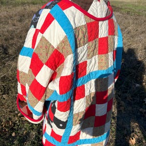 May include: A quilted jacket featuring a patchwork design with red, white, blue, and tan squares. The jacket has a red trim and is worn by a person. The jacket is a unique piece of wearable art.