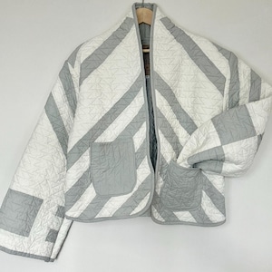May include: A quilted jacket in a white and gray geometric pattern. The jacket has two front pockets and a relaxed fit. The jacket is hanging on a wooden hanger against a white background.