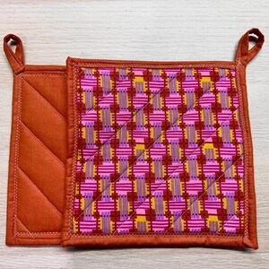 May include: Two quilted pot holders. One is solid rust orange with diamond quilting. The other is rust orange with a pink, purple, and yellow geometric pattern.