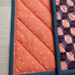 May include: Two quilted pot holders with blue denim trim. One pot holder has a peach background with white polka dots. The other pot holder has a black, white, and pink geometric pattern.