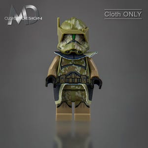 May include: A green and brown camouflage patterned Lego minifigure with a green helmet and a blue cape. The minifigure is wearing a brown belt and has a green face.