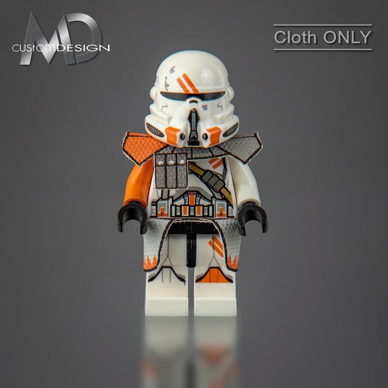 May include: A white and orange Lego minifigure of a Star Wars clone trooper with a helmet and a blaster rifle. The figure has a black and orange chest piece and a black and orange leg piece. The figure is standing on a gray surface. "Cloth ONLY" is written in white text on a black background in the upper right corner of the image.