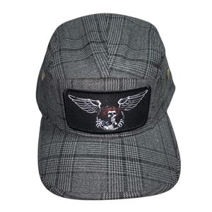 May include: A five-panel cap in a grey and black plaid pattern. The front features a rectangular black patch with a white graphic of a skull with wings and a red circular accent. The cap has a flat brim and metallic side eyelets for ventilation.