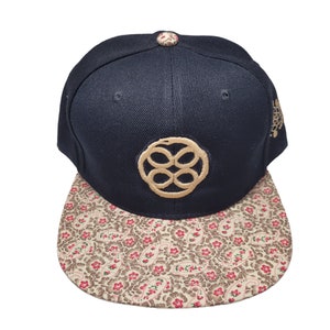 Könnte beinhalten: Eine schwarze und goldene Baseballkappe mit einem floralen Muster am Schirm. Die Kappe hat ein golden gesticktes Logo mit einem Kreisdesign.