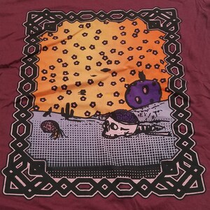 May include: A maroon t-shirt with a black and pink patterned frame. Inside the frame is a colorful illustration of a skull, a purple planet, and a black cat. The background is a yellow and orange gradient with black dots.