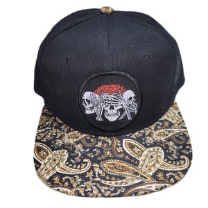 May include: A black baseball cap featuring a circular patch on the front panel with three white skulls and red roses. The skulls are positioned to mimic the see no evil, hear no evil, speak no evil gesture. The flat brim has a black and gold paisley pattern.