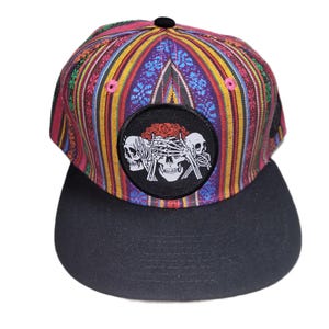 May include: A baseball cap featuring a vibrant, multi-colored woven pattern with vertical stripes in shades of purple, red, orange, and green. The front panel displays a circular black patch with a white graphic of three skulls.