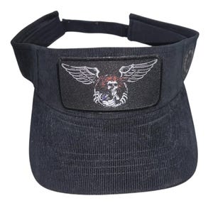 May include: A black corduroy visor featuring a rectangular patch on the front. The patch displays a graphic of a skull with wings, rendered in white, red, and blue tones. The visor has an adjustable strap at the back for a secure fit.