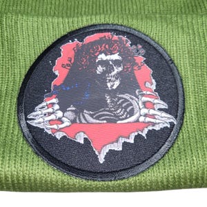 May include: Olive green beanie featuring a circular patch with a skull design. The patch has a black border, a red background, and a detailed illustration of a skeleton with outstretched hands. The beanie is made of a knitted fabric.