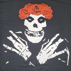 May include: Black t-shirt featuring a white skull with a red rose crown and skeleton hands. The graphic design is a stylized illustration. The shirt is a dark color and has a classic crew neck.