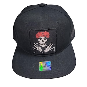 May include: Black baseball cap with a flat brim. The cap features a rectangular patch with a skull design wearing a red rose crown. A holographic sticker is on the brim. The cap is a size 7 1/8.