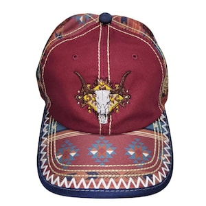 May include: A maroon baseball cap featuring a central embroidered white bull skull graphic with dark horns. The cap includes a patterned brim and side panels with geometric shapes in blue, orange, and white, finished with white zigzag stitching.