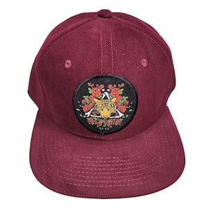 May include: A burgundy corduroy baseball cap with a curved brim. The cap features a circular patch with a tiger design inside a triangle, surrounded by roses and leaves.