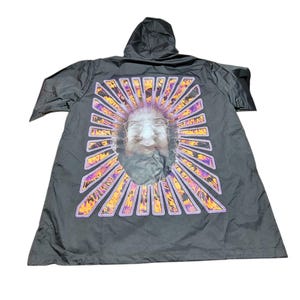 May include: Black hooded jacket featuring a graphic of a face within a sunburst design. The sunburst has orange, purple, and yellow rays. The jacket has a hood and is made of a lightweight material.