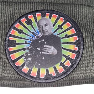 May include: A dark green beanie featuring a circular patch with a black and white image of a man holding a molecular model. The patch has a colorful, rainbow-like design radiating outwards.