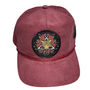 May include: Maroon corduroy baseball cap with a round patch featuring a tiger, roses, and a triangle design. The cap has a curved brim and a red rope detail. A stylish accessory.